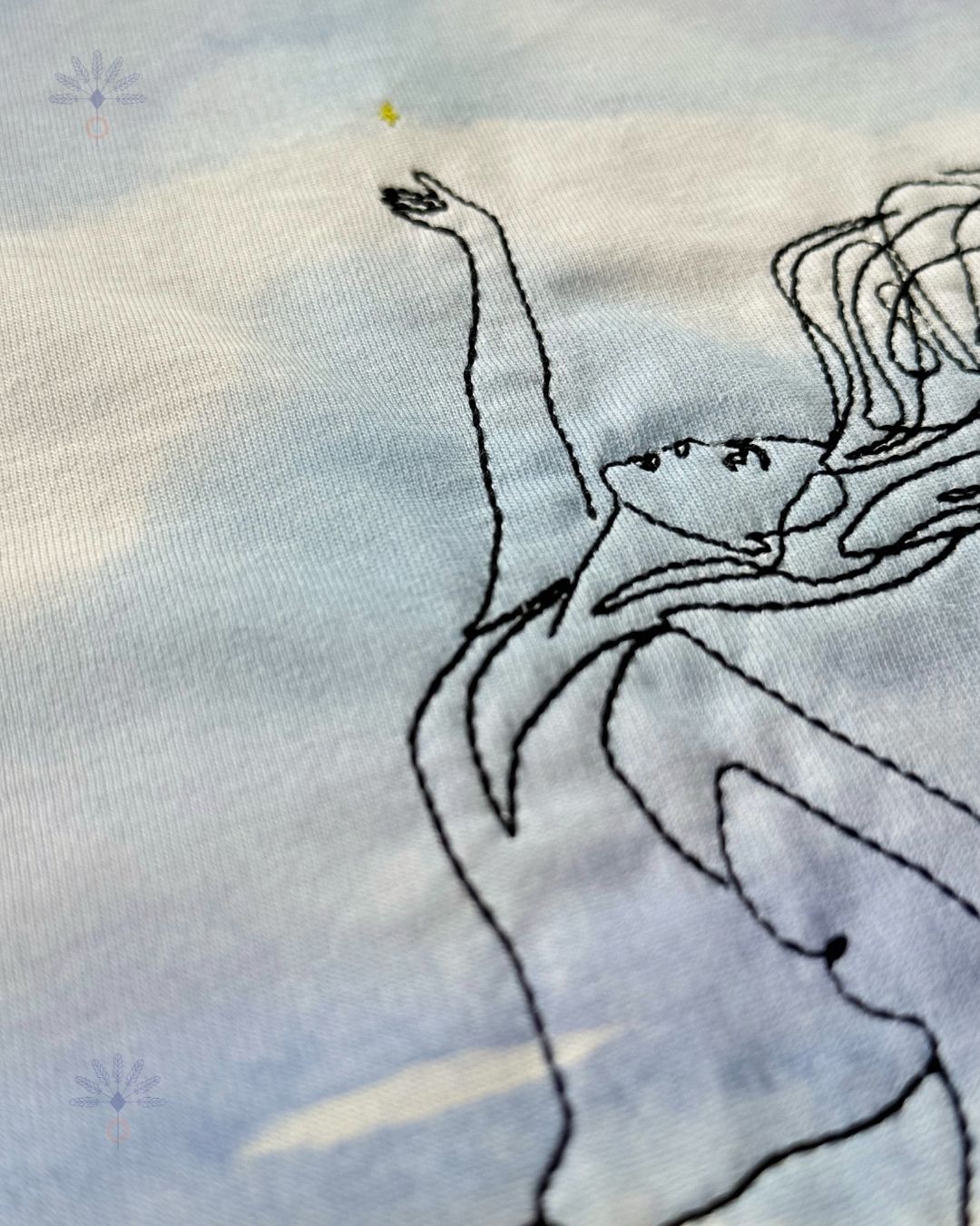 Détail de la broderie silhouette en ligne noire sur le tee-shirt Soamarena AMA, avec motif imprimé bleu sur coton biologique blanc.