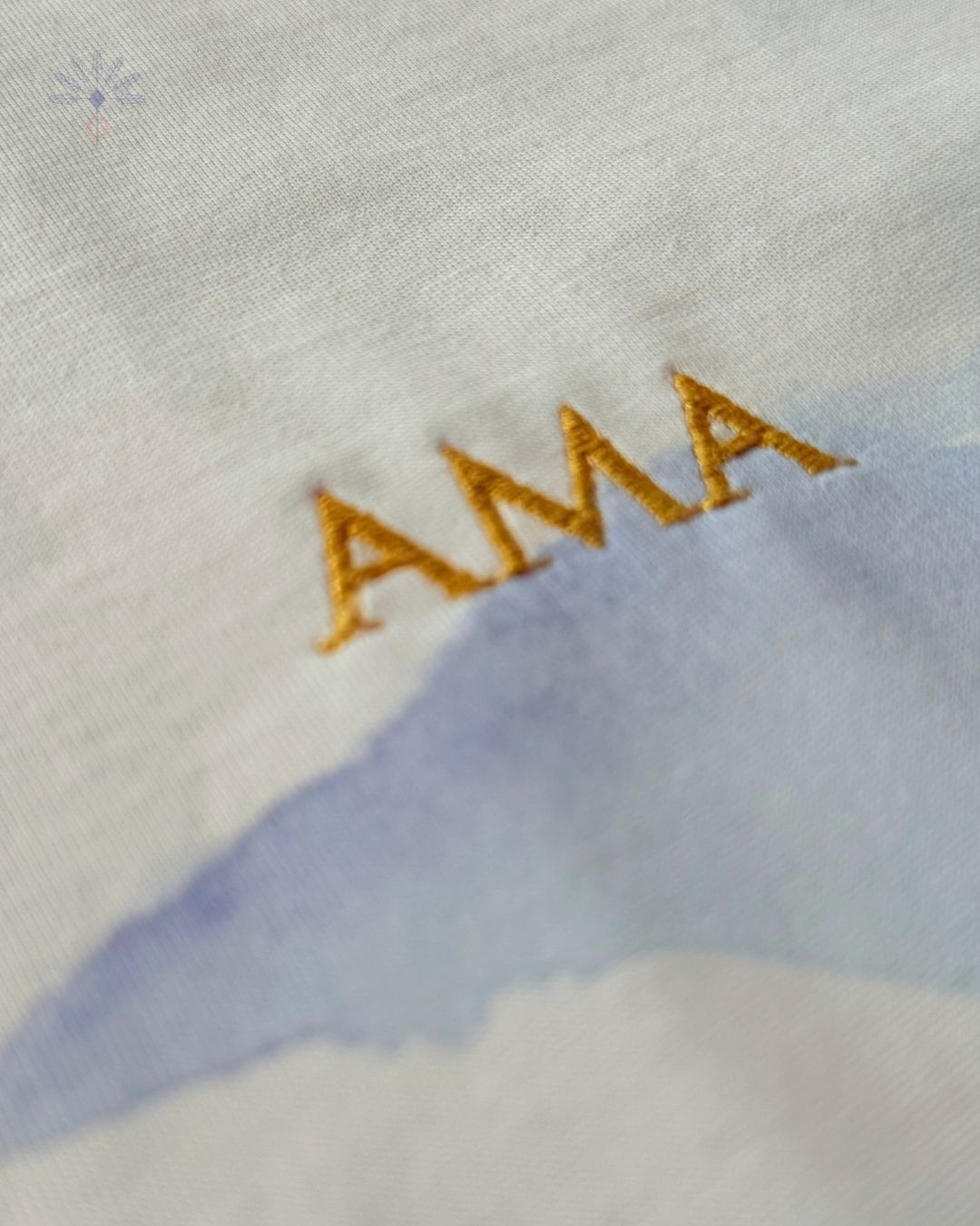 Détail de la broderie dorée du texte « AMA » sur le tee-shirt Soamarena, en coton biologique blanc avec motif imprimé bleu.