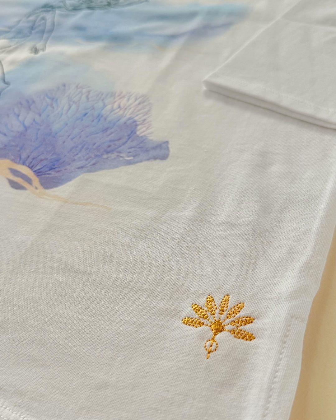 Signature de la broderie dorée sur le tee-shirt Soamarena AMA, en coton biologique blanc, avec motif artistique bleu imprimé en arrière-plan.