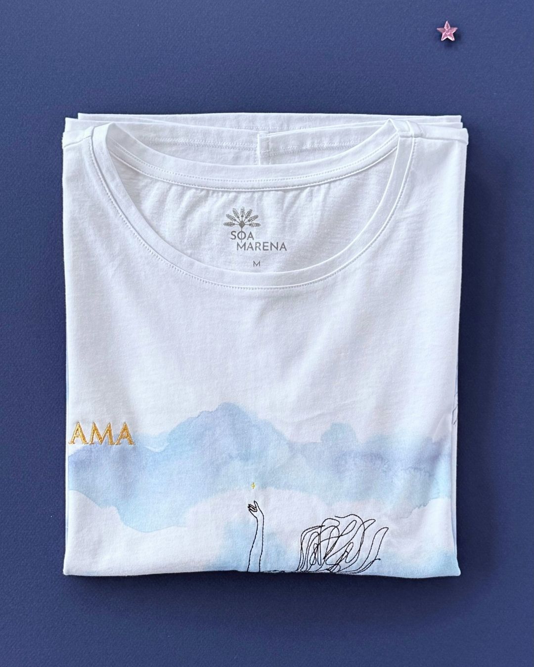 Tee-shirt Soamarena AMA plié, en coton biologique blanc, avec détail de broderie étoile près de l’encolure et motif artistique visible.